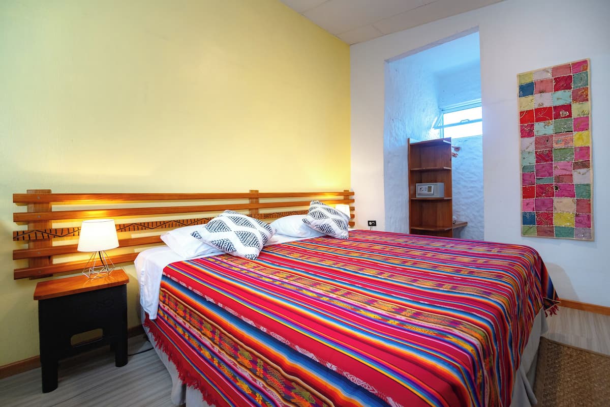 A bedroom with a wooden headboard, a colorful bedspread, a bedside lamp, and a quilted wall decoration.