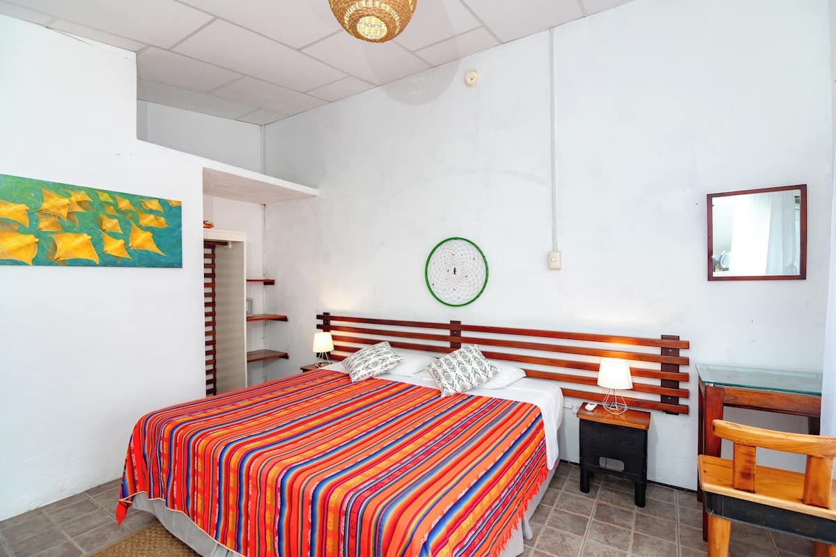 A bedroom with a bed, a bedside table with a lamp, a chair, a wall clock, and a mirror.