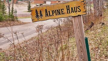 A wooden sign with 'Alpine Haus' written on it, surrounded by trees and a road.
