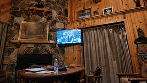 Una habitación acogedora con pared de piedra, paneles de madera, un ciervo montado en la pared, televisión, chimenea y una mesa de comedor con sillas.