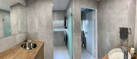 Un baño moderno con un mueble de tocador de madera, un lavamanos de acero inoxidable, un cerramiento de ducha de vidrio y un inodoro blanco.