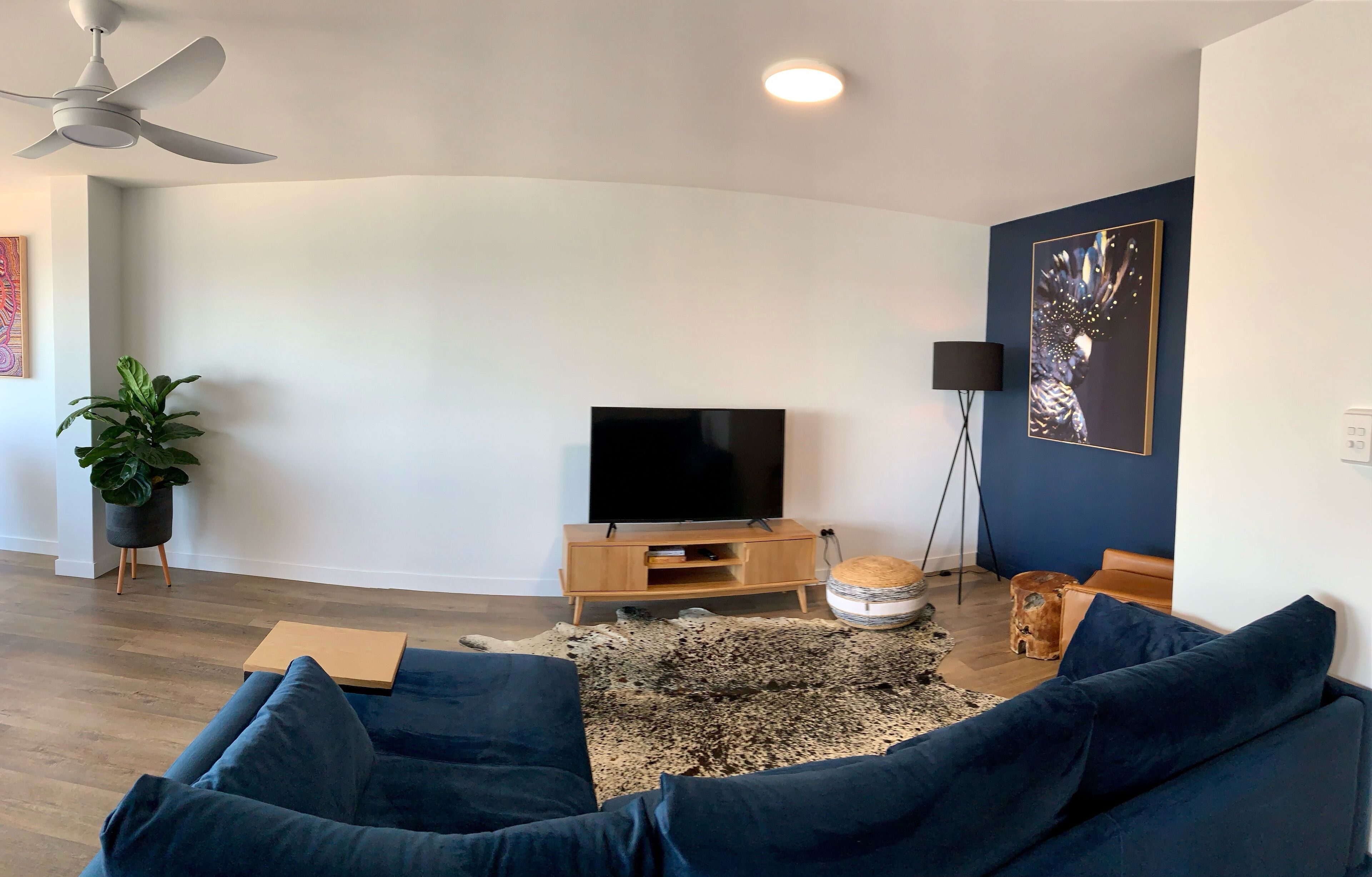 Una sala de estar moderna con un televisor de pantalla plana, un ventilador de techo, una planta en maceta, un sofá azul, un tapete y una obra de arte en la pared.