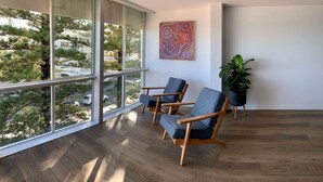 Une pièce moderne avec de grandes fenêtres, deux chaises en bois, une plante en pot et un tableau abstrait aux couleurs vives.