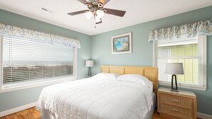 Un dormitorio con una cama, dos mesitas de noche, un ventilador de techo y dos ventanas con persianas.