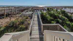 Une promenade en bois mène à une plage où se trouvent des chaises blanches et une piscine.