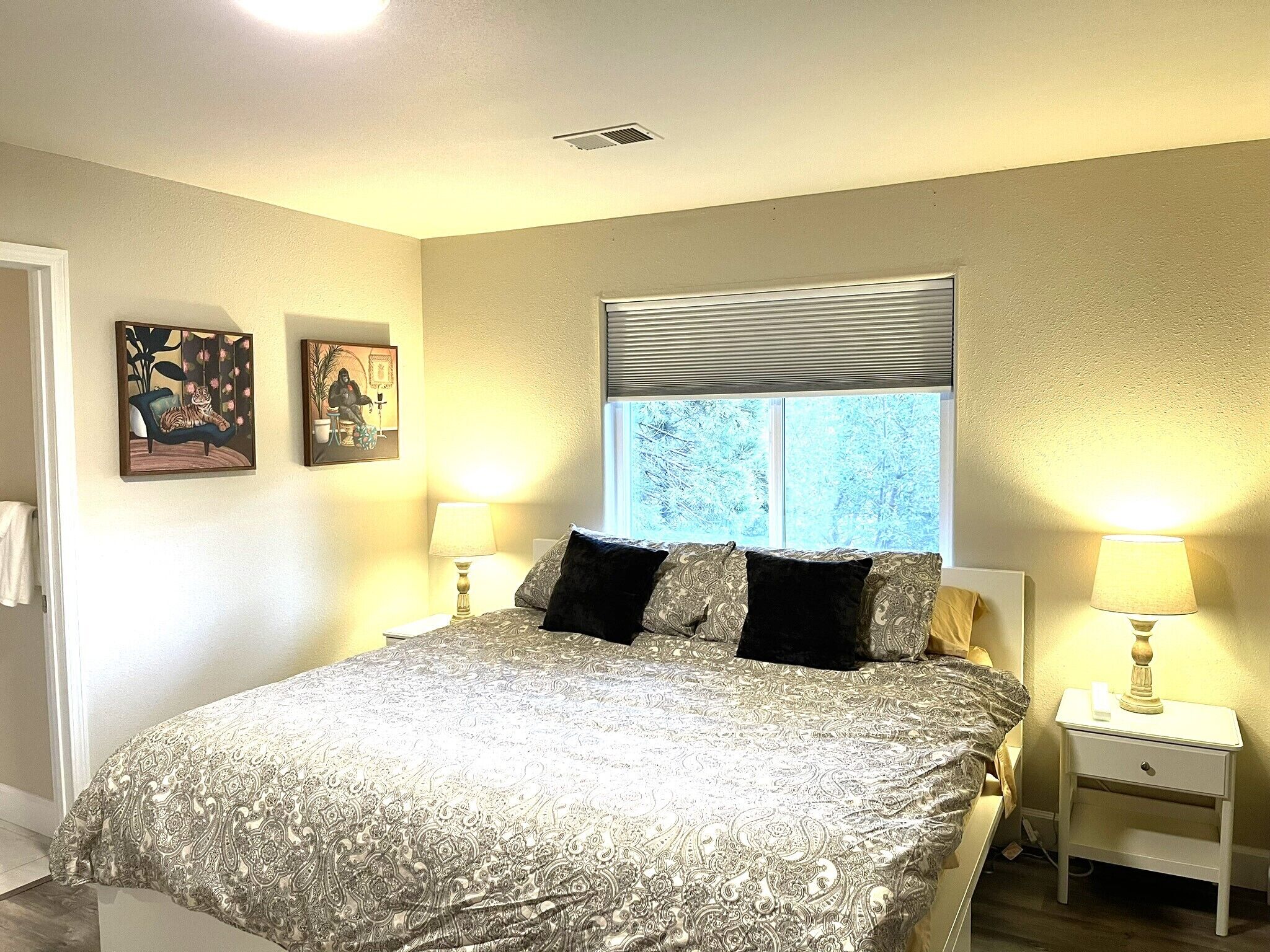 A bedroom with a bed, two nightstands, a window with blinds, and two wall-mounted paintings.