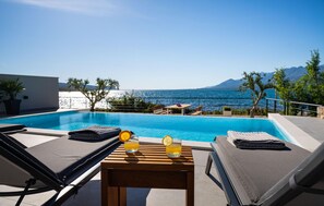 Ein Pool auf dem Dach mit Liegestühlen, einem Tisch mit Getränken und Blick auf das Meer und die Berge.