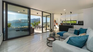 A modern living room with a large sliding glass door leading to a balcony overlooking a body of water and mountains. - Lovely apartment in Rovanjska (Rovanjska)