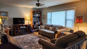 A living room with a sofa, a television, a bookshelf, and a large window. - 4 minutes from Saratoga Racetrack, this home is clean & cozy, with private deck (Saratoga Springs)
