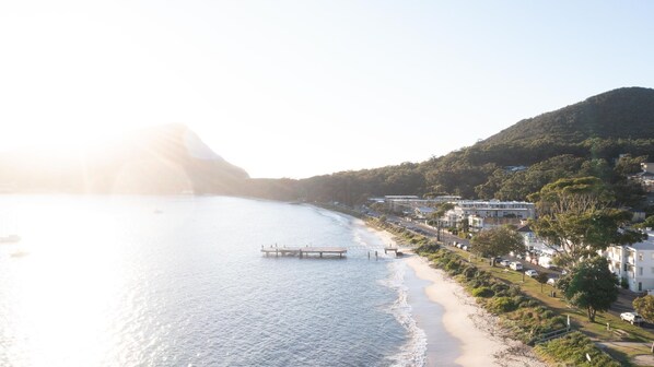 A coastal area with a pier extending into calm waters, surrounded by residential buildings and a hilly backdrop.