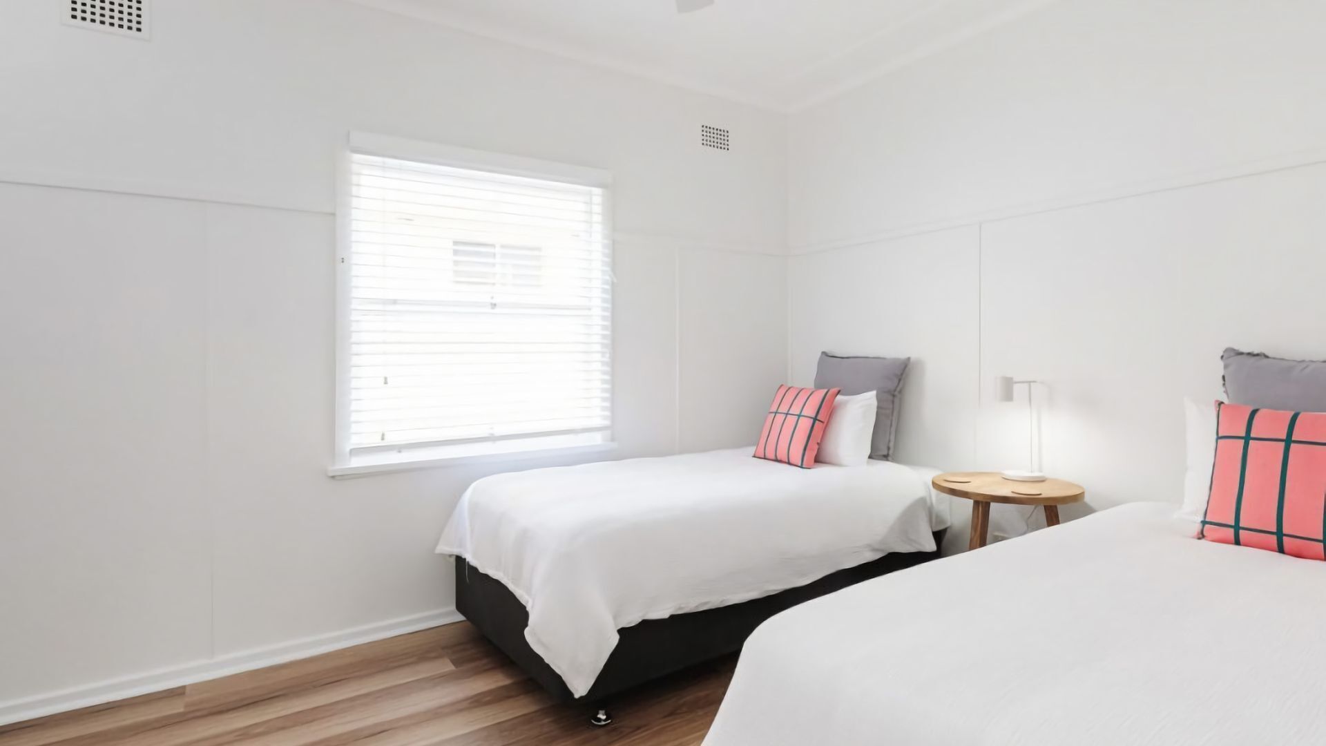 Un dormitorio limpio y minimalista con dos camas, una mesita de noche pequeña y una ventana con persianas.