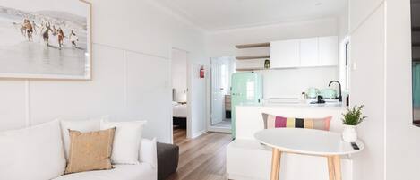 Un salón moderno y bien iluminado con un sofá blanco, una pequeña mesa redonda y una cocina al fondo.