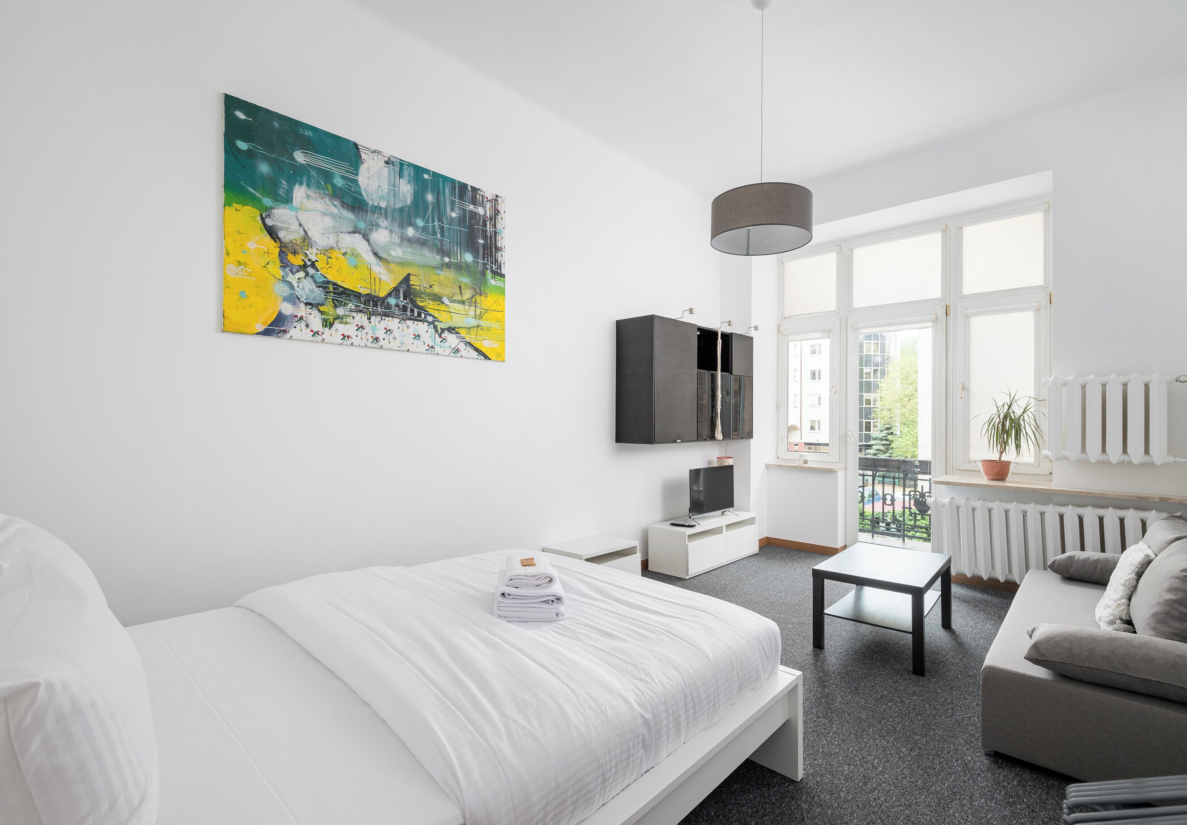 A modern hotel room with a large bed, a flat-screen TV, a small table, a sofa, and a balcony with a view of the city.