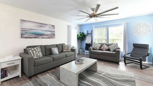 A living room with two sofas, a coffee table, a bookshelf, a ceiling fan, and a large window with blinds. - Green Fairways Retreat Condo (Myrtle Beach)