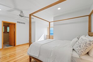 Een slaapkamer met een hemelbed, houten balken, een plafondventilator en uitzicht op de zee door een raam.