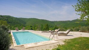 Ein rechteckiger Pool mit klarem, blauem Wasser, umgeben von einer Kiesfläche mit Liegestühlen und Blick auf grüne Hügel im Hintergrund.