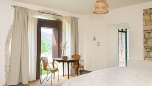 Ein Schlafzimmer mit Bett, zwei Stühlen, einem Tisch mit einer Vase, einer Hängelampe und Blick auf Grün durch das Fenster.