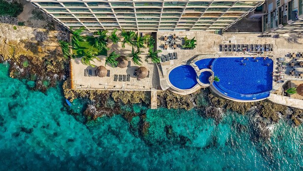 An aerial view of a resort with a large pool area, palm trees, and a rocky shoreline.