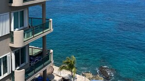 A multi-story building with balconies overlooking a clear blue sea, a small boat in the distance, and a rocky shoreline.