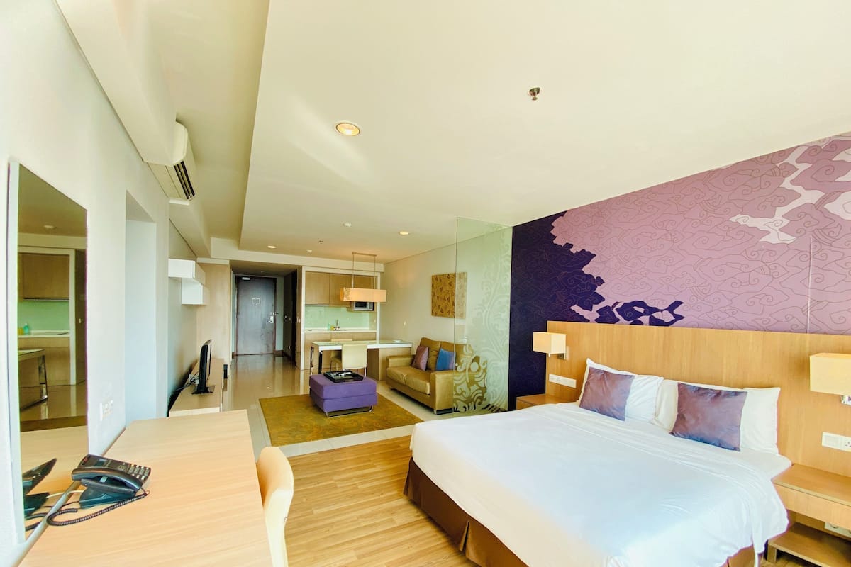 A modern hotel room with a large bed, a desk, a sofa, and a kitchen area visible through an open door.