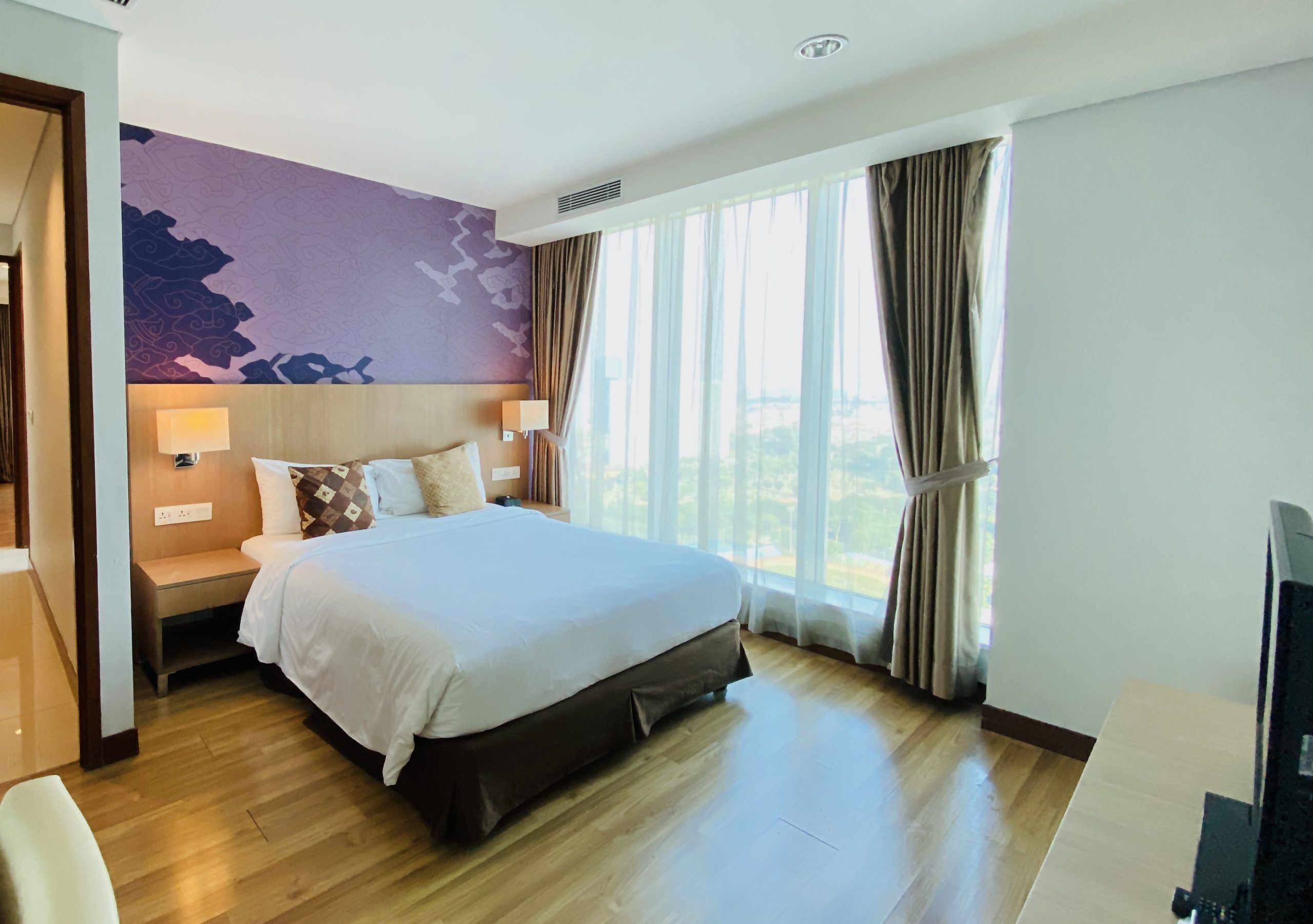 A hotel room with a large bed, a nightstand with a lamp, a view of the outdoors through large windows, and a modern wall design.
