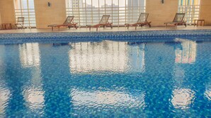 Piscina interna com água azul cristalina, rodeada por espreguiçadeiras e grandes janelas que oferecem vista para prédios.