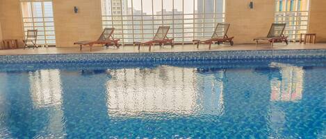 An indoor swimming pool with clear blue water, surrounded by lounge chairs and large windows offering views of buildings.