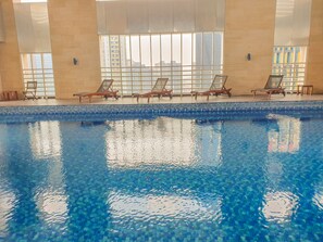 Piscina interna com água azul cristalina, rodeada por espreguiçadeiras e grandes janelas que oferecem vista para prédios.