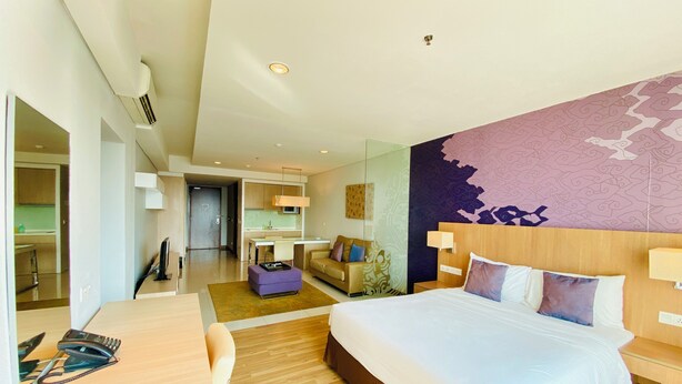 Quarto de hotel moderno com uma cama grande, uma mesa de escritório, um sofá e uma área de cozinha visível através de uma porta aberta.