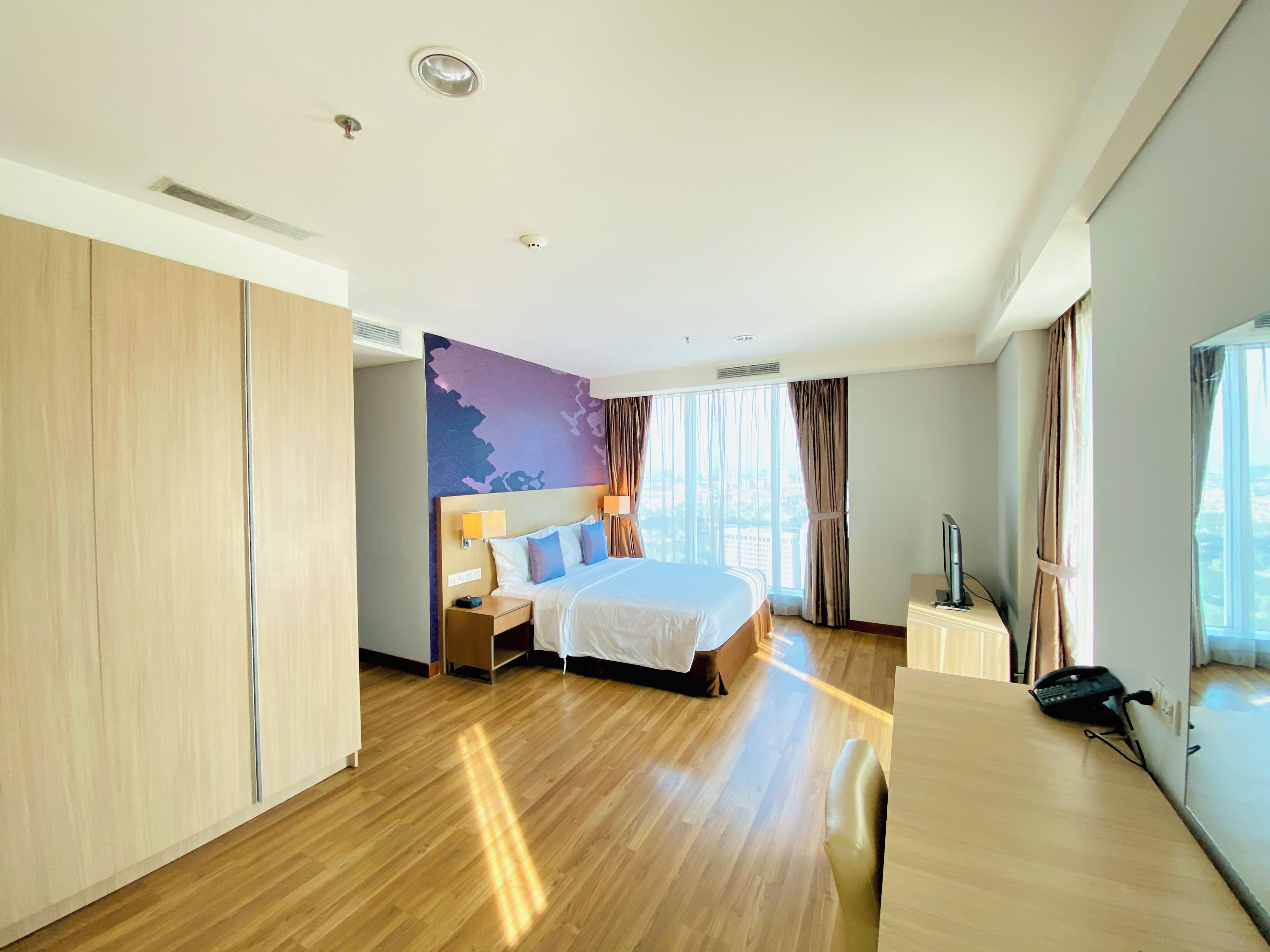 A modern hotel room with a large bed, wooden flooring, a desk with a telephone, and a view of the city through large windows.