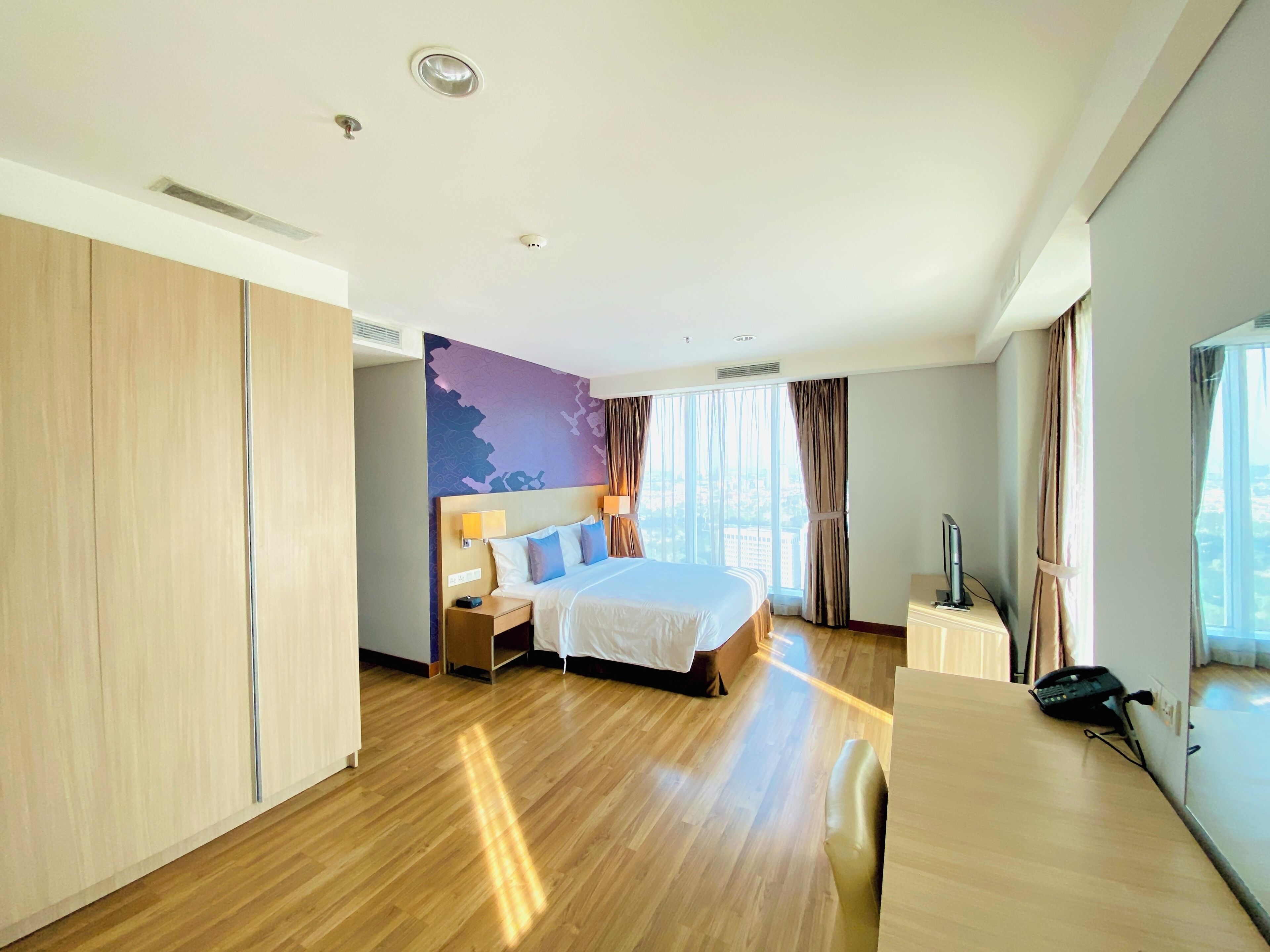 A modern hotel room with a large bed, wooden flooring, a desk with a telephone, and a view of the city through large windows.