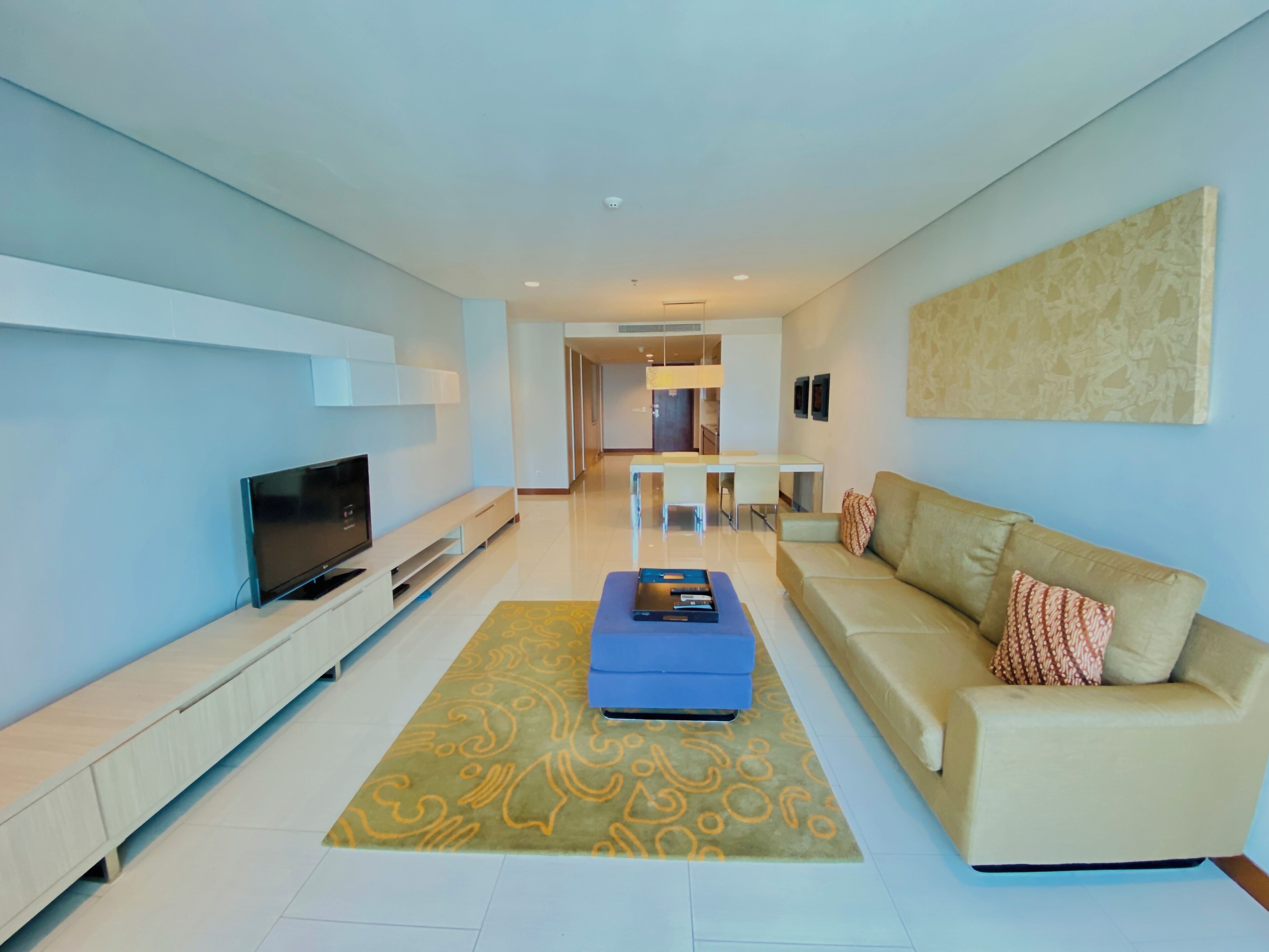 A modern living room with a flat-screen TV, a beige sofa, a blue ottoman, and a patterned rug.