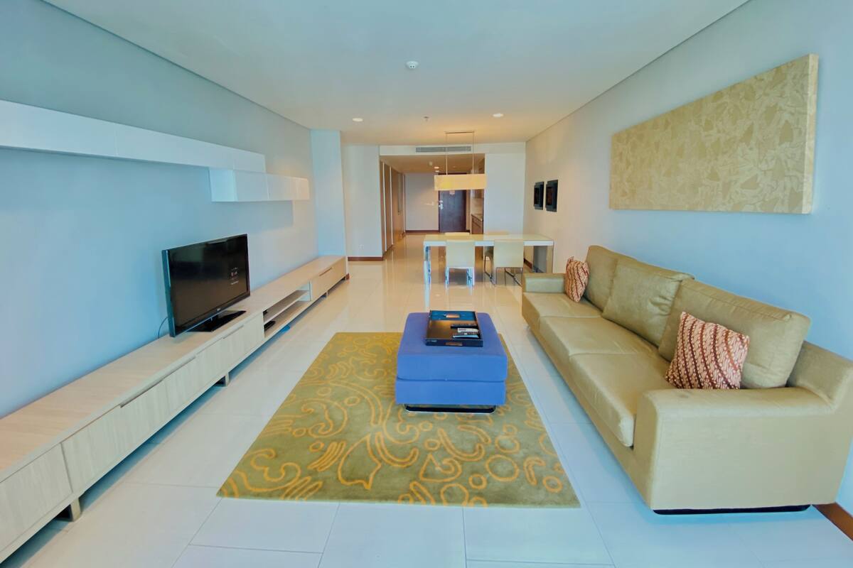 A modern living room with a flat-screen TV, a beige sofa, a blue ottoman, and a patterned rug.