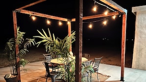 An outdoor patio area with a wooden pergola, string lights, a table with chairs, and potted plants.