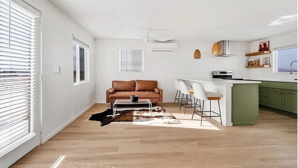 A modern living room with a brown leather sofa, a coffee table, and a cowhide rug. There is a kitchen area with a white countertop and green cabinets.
