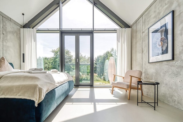 A modern bedroom with a large bed, a chair, a small table, and a balcony with a view of greenery.