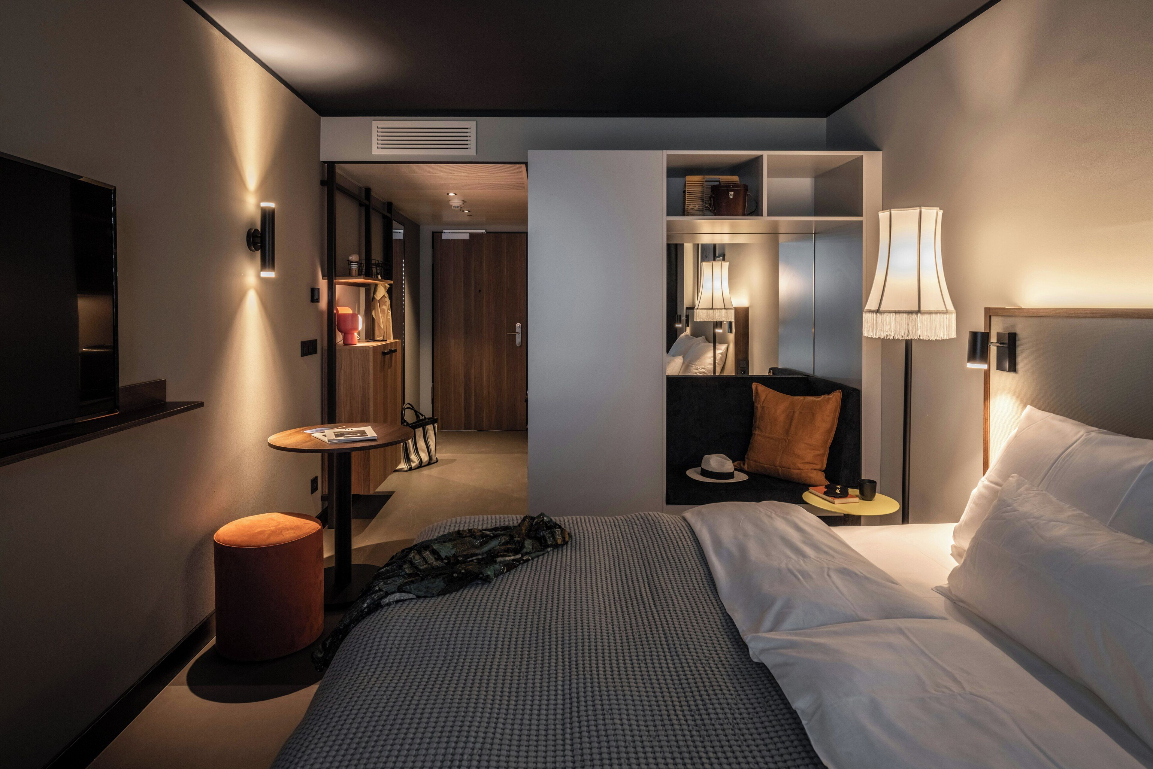 A hotel room with a bed, bedside table, lamp, and a small table with a hat and cup.