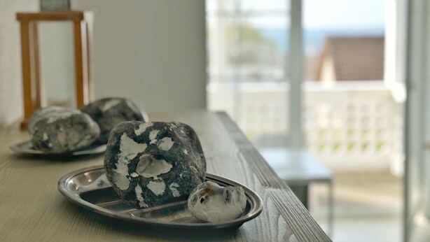 A wooden table with three stone sculptures and a small wooden stool in a bright room with a view of the sea.