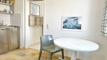 A compact kitchen with a dining area, featuring a round table and two chairs, a refrigerator, and a shelf with kitchenware.