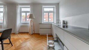 A modern kitchen with white cabinets, a wooden floor, and a view of a red-roofed building through the windows.