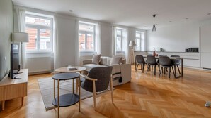 A modern living room with a sofa, coffee table, and chairs, a dining area with a table and chairs, and a kitchen in the background.