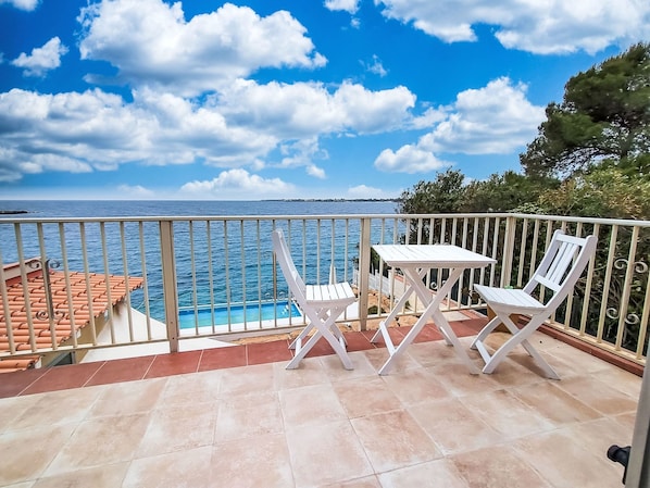 A balcony with a pool, white chairs, and a view of the sea.