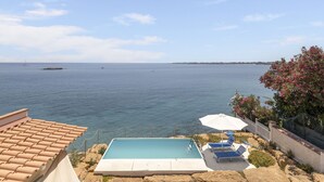 A swimming pool with a view of the sea, a white fence, and a pink flowering tree. - Norbert by Interhome (Siracusa)