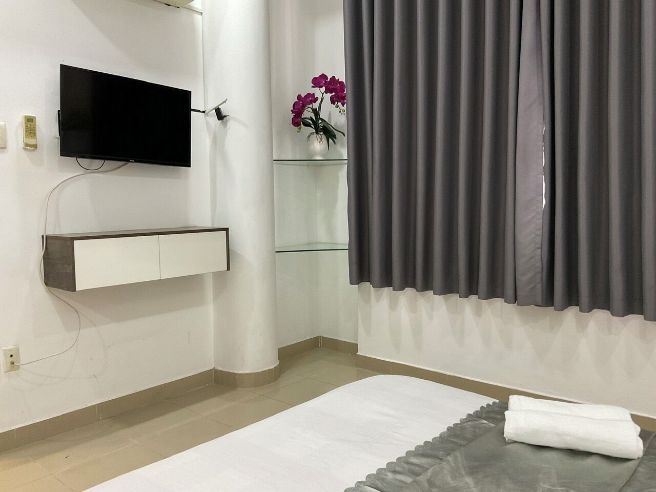A hotel room with a flat-screen TV mounted on the wall, a glass shelf with a vase of flowers, grey curtains, a bed with a white mattress and grey blanket, and a white bedside table.