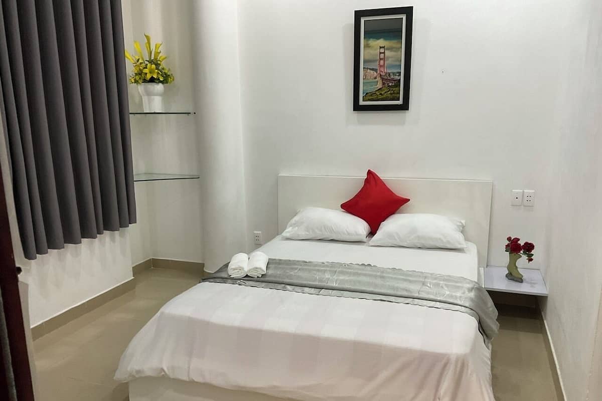 Ein ordentlich gemachtes Bett mit weißer Bettwäsche und einem roten Kissen, ein kleiner Nachttisch mit Blumen in einer Vase und ein gerahmtes Bild an der Wand.