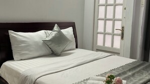 A neatly made bed with white linens and pillows, a decorative pillow with a floral design, and a rolled towel with a pink flower on a bed skirt. - Minh Chau Hotel - Dien Bien Phu (Ho Chi Minh City)