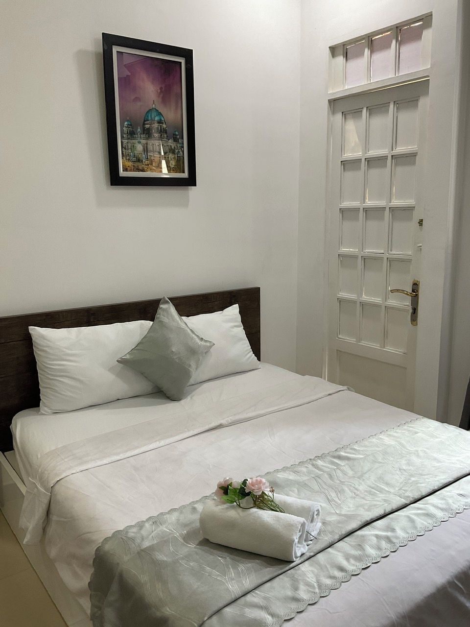 A neatly made bed with white linens and a pillow, a framed artwork above it, and a white door with a window to the right.