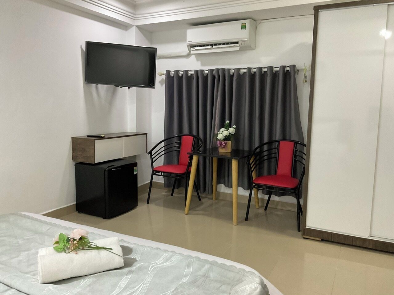A hotel room with a bed, a TV, a small desk, two chairs, a table with flowers, and a curtain.