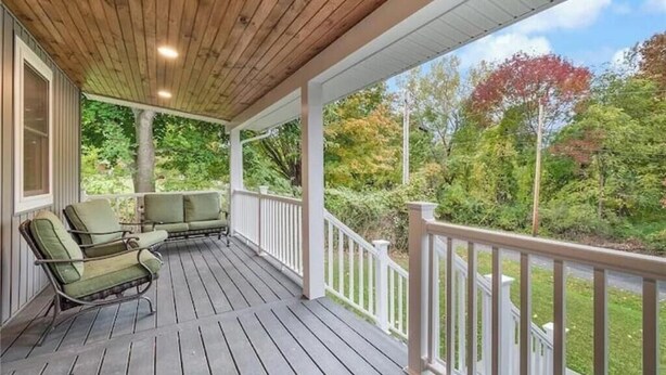 Eine überdachte Veranda mit Holzboden und weißen Geländern, ausgestattet mit grünen, gepolsterten Sesseln und Blick ins bewaldete Gebiet.