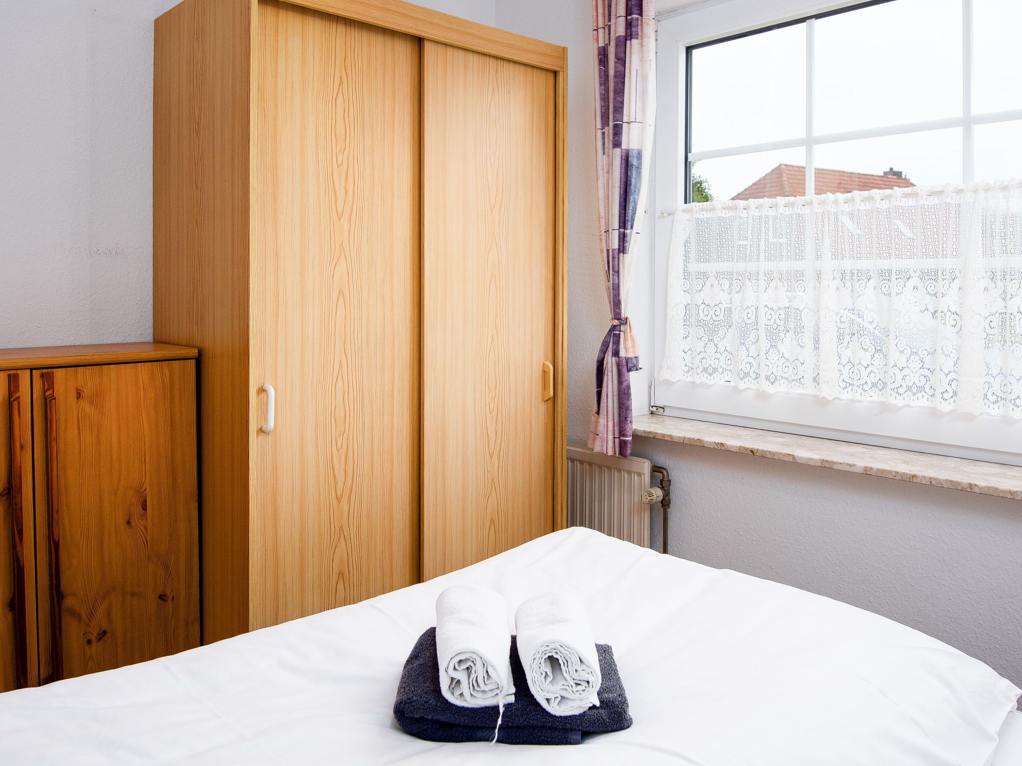Une chambre à coucher avec une armoire en bois, un lit recouvert de serviettes blanches et bleu foncé, et une fenêtre ornée de rideaux en dentelle.
